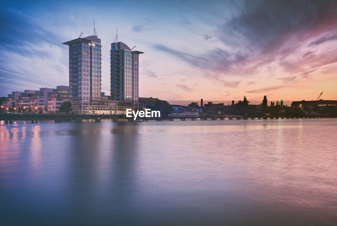 Cityscape by spree river against sky during sunset