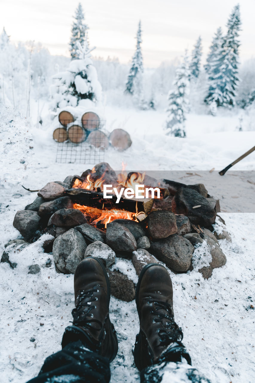 Crop anonymous traveler in warm boots resting near bonfire on snowy ground near coniferous forest in winter person