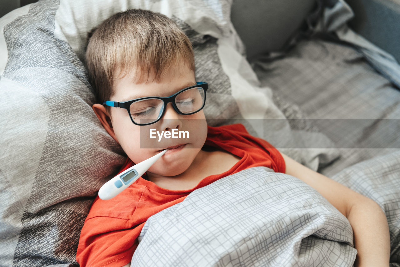 A boy with glasses lies on a bed with a thermometer in his mouth