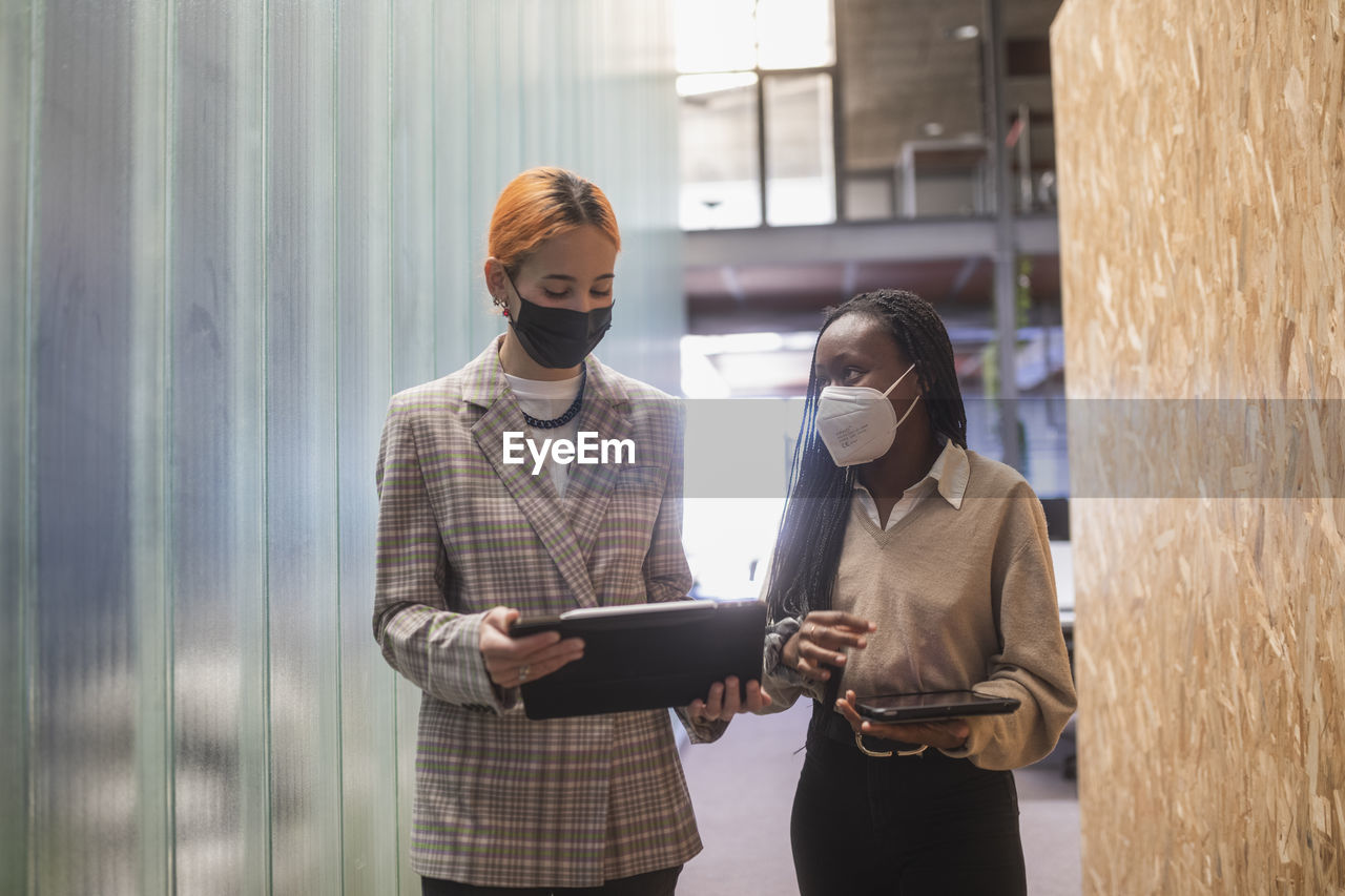 Multiethnic female colleagues with tablets and in protective masks standing in coworking space and discussing project