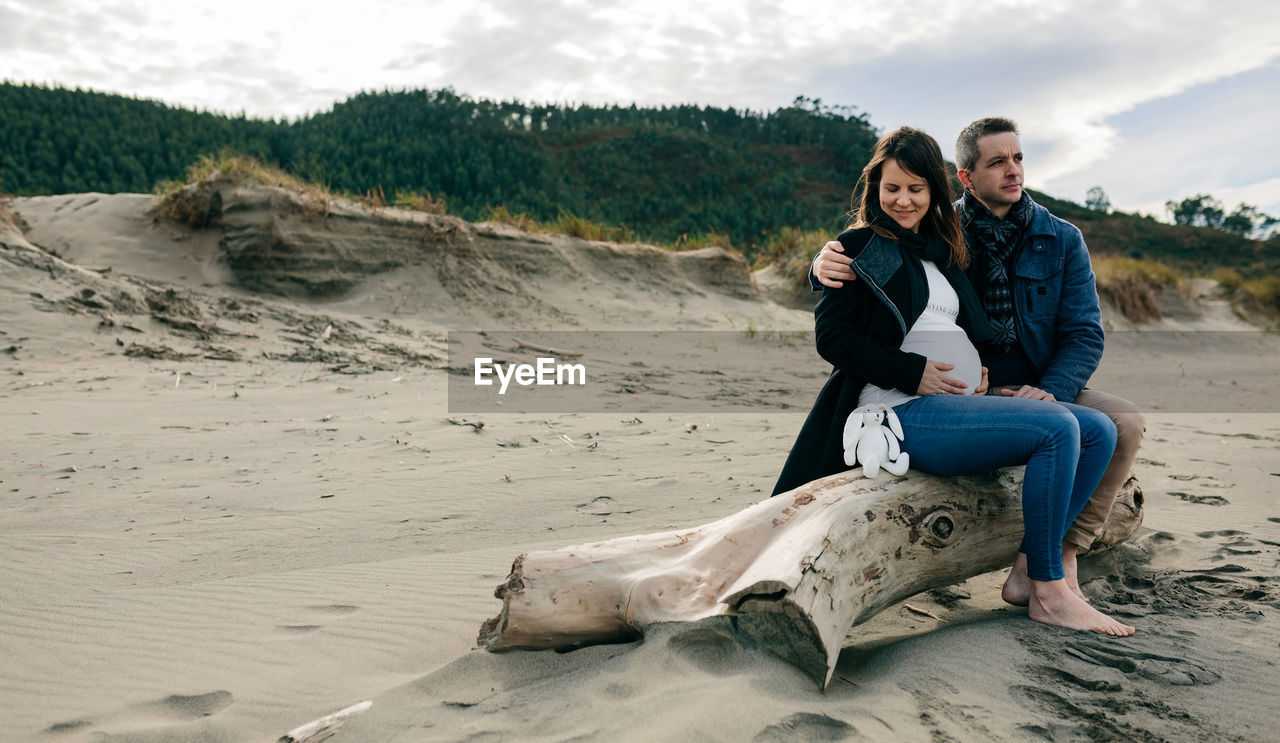 Pregnant woman sitting with man on wood at beach