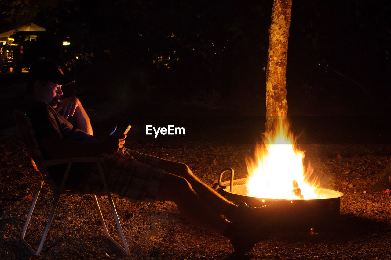 Man using mobile phone while sitting by fire pit in back yard at night