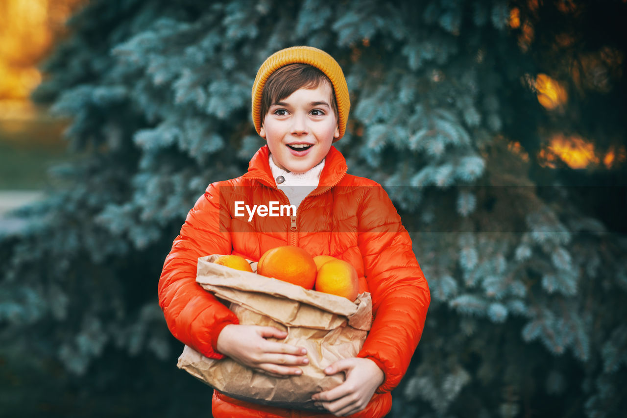 A boy in a bright orange jacket and a yellow hat holds a large package with oranges 