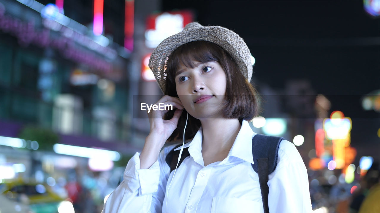 Woman listening music through in-ear headphones while standing in city at night