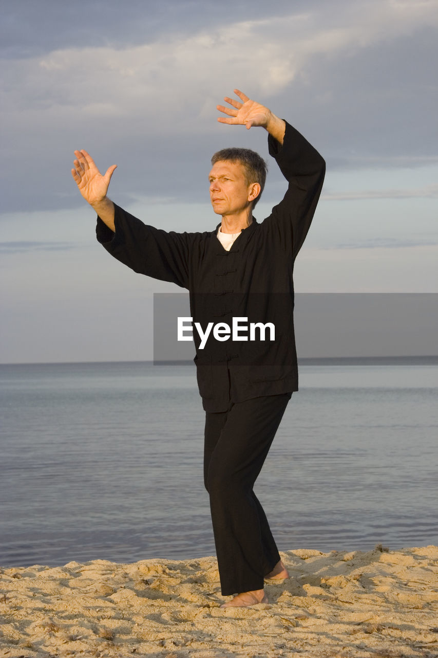 Full length of mature man practicing tai chi on sea shore at beach against cloudy sky