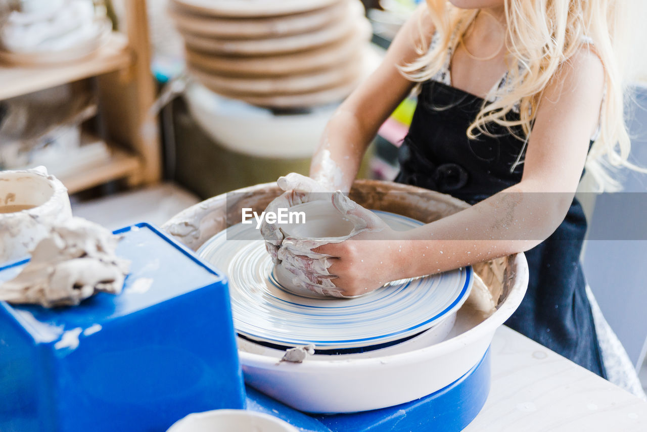 Crop unrecognizable little girl in black apron standing near pottery wheel while shaping clay pot in light workshop