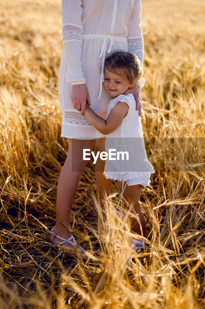 Daughter hugging mother's leg on yellow wheat field evening in summer