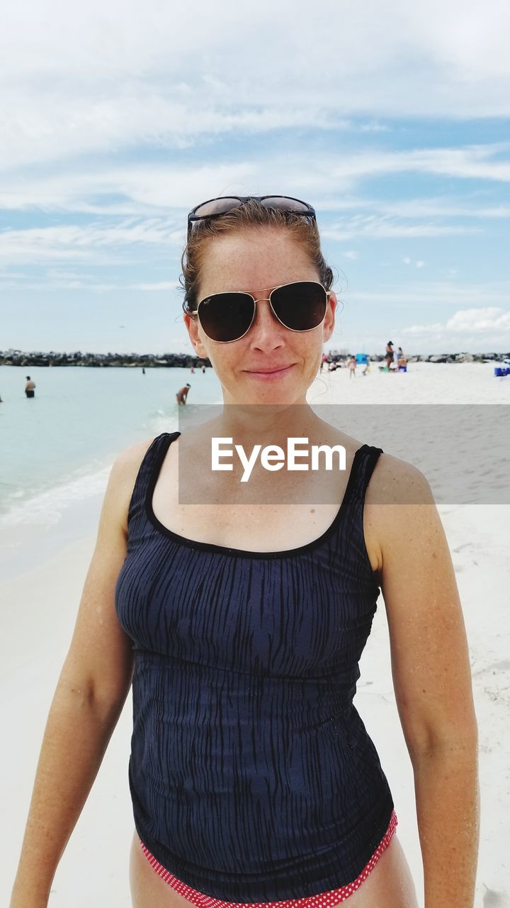 Portrait of woman wearing sunglasses standing at beach against sky