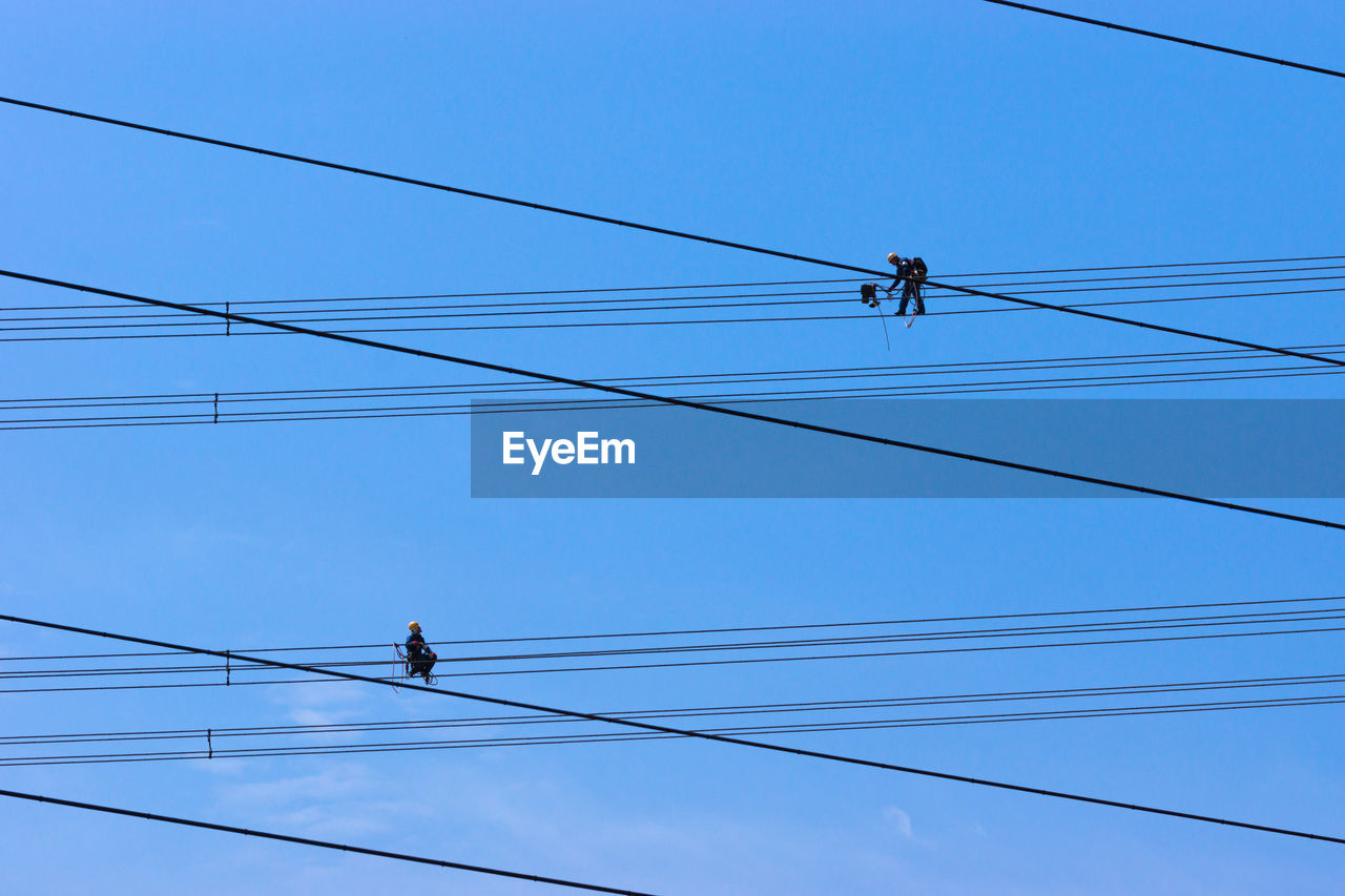 Low angle view of workers working on electricity pylon cables against clear blue sky