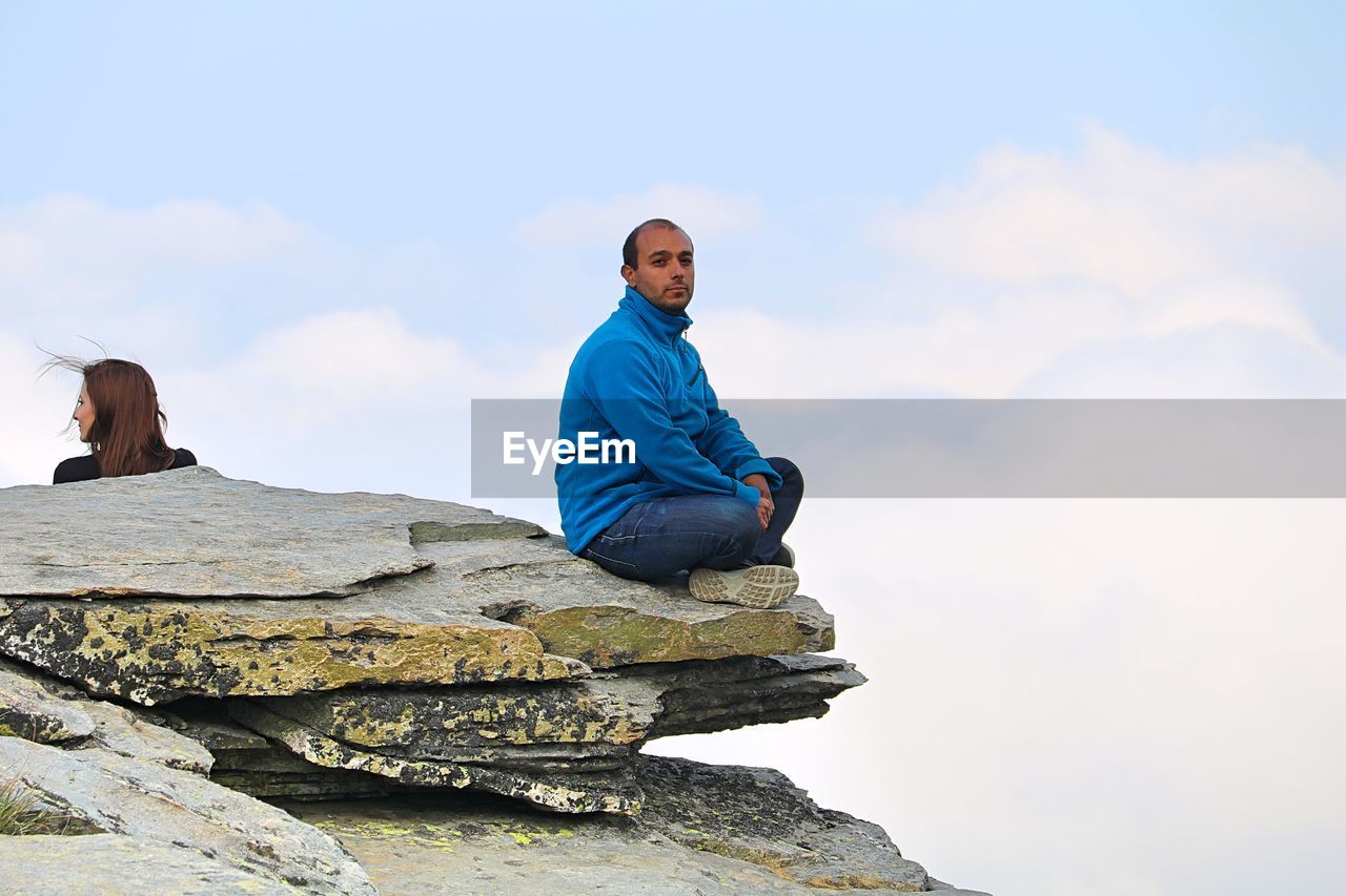 Young man sitting on rock against sky