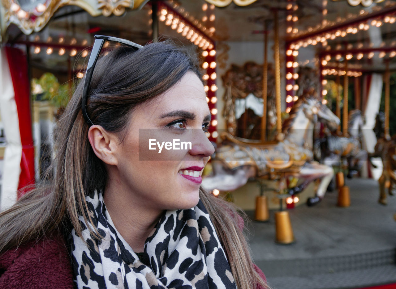 PORTRAIT OF SMILING YOUNG WOMAN WITH CAROUSEL IN AMUSEMENT PARK