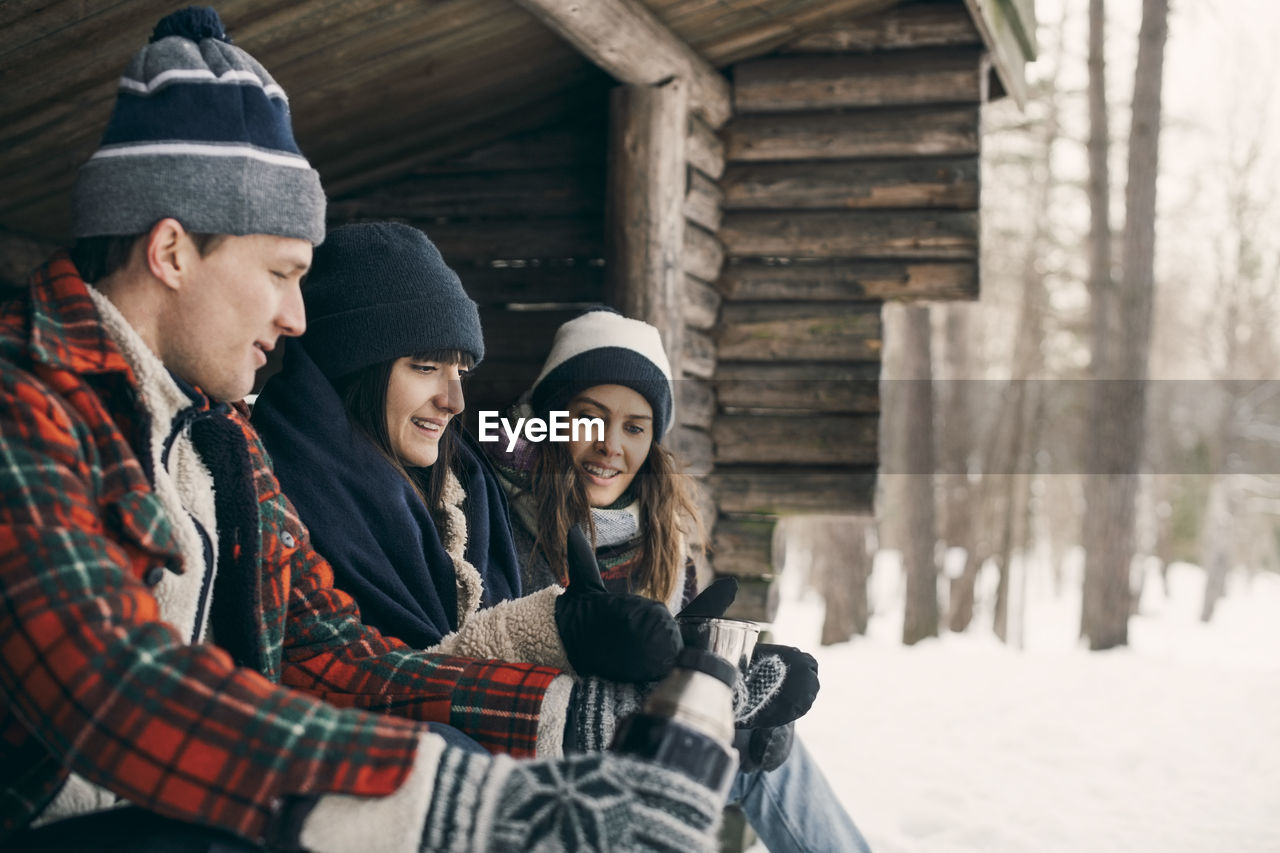 Man giving drink to female friends while sitting at log cabin during winter