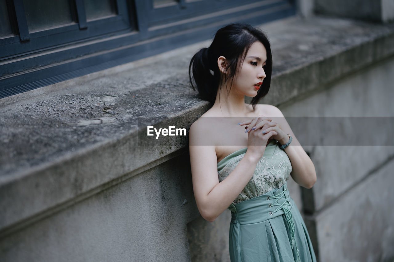 young woman looking away while standing against wall