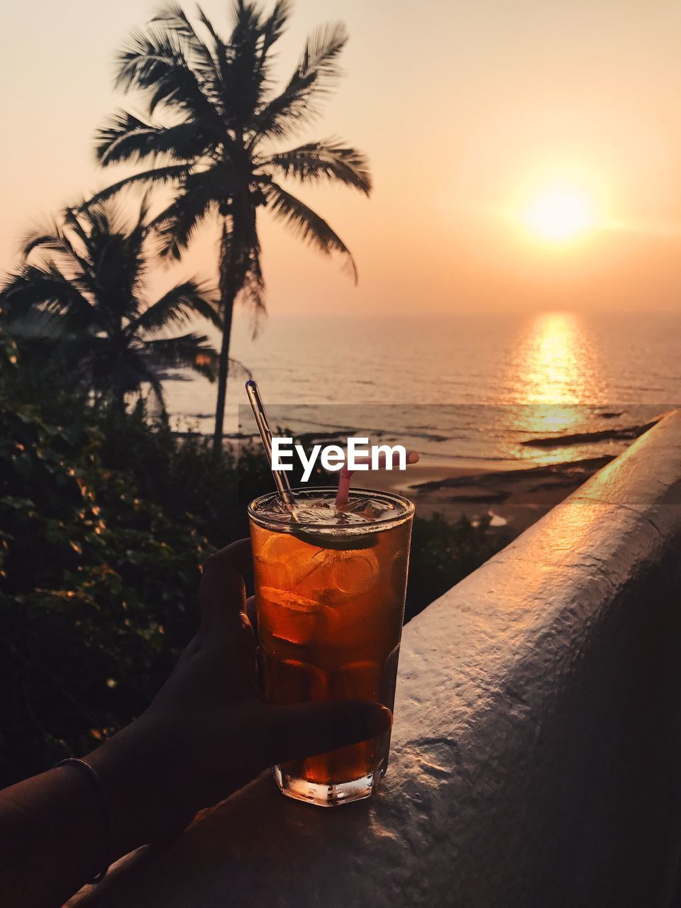 Cropped image of hand holding drink against sea during sunset