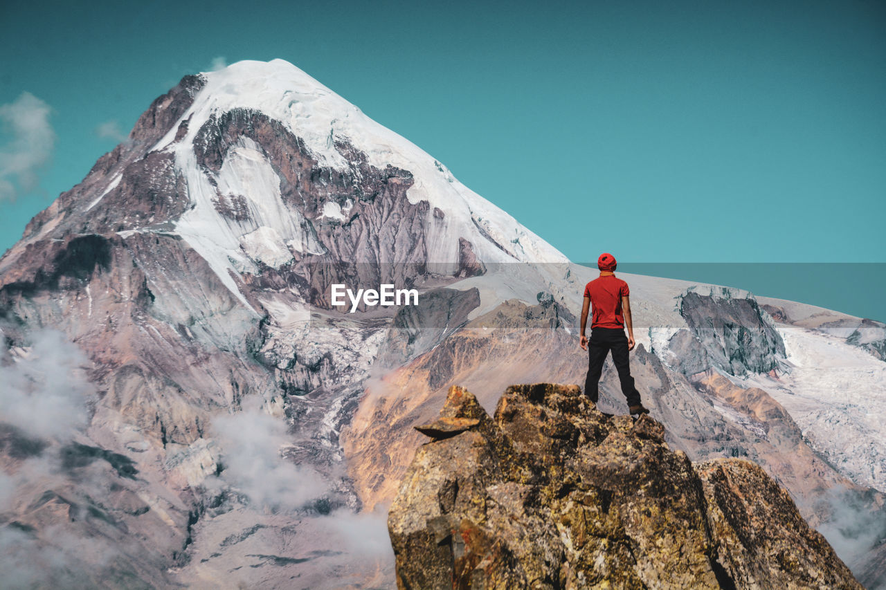 Rear view of young man standing on rock formation against mountain and sky