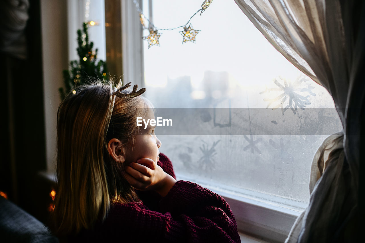 Side view of girl looking through window at home in christmas time
