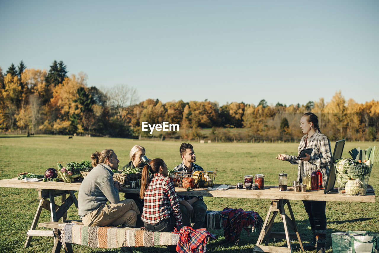 Instructor holding digital tablet while talking with farmers sitting at table on field