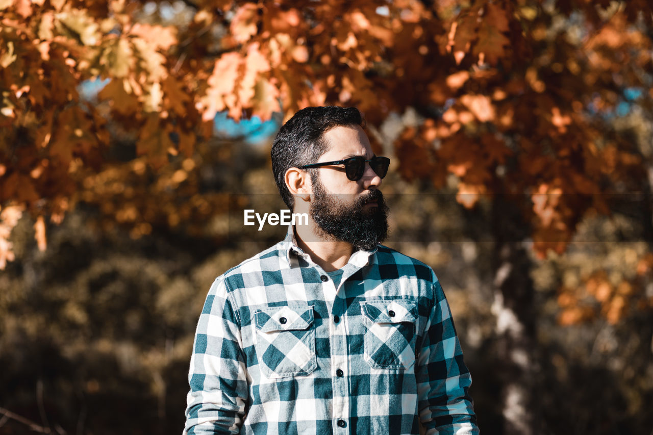 Bearded man looking away while wearing sunglasses against trees