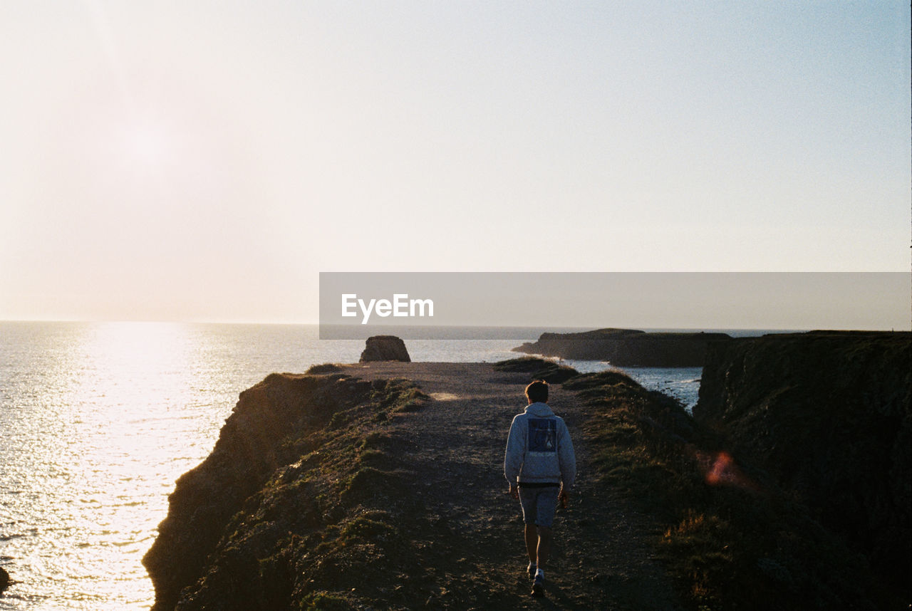 Rear view of man walking on cliff at sea against clear sky during sunset