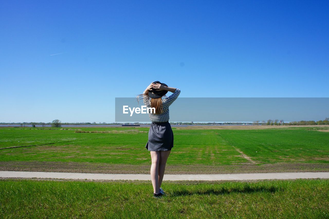 Rear view of woman wearing hat standing on grassy field against sky during sunny day