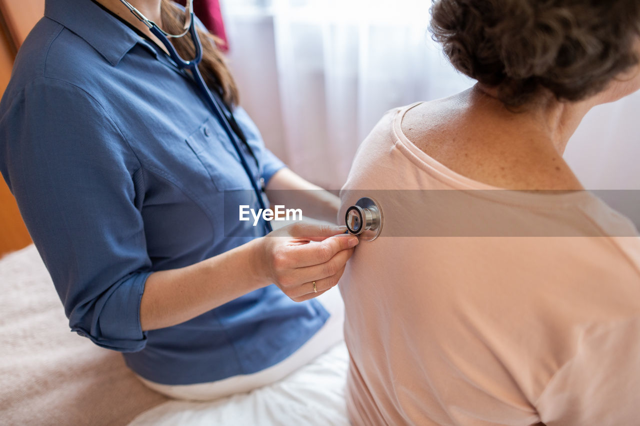 Cropped view of nurse putting stethoscope on back of elderly female patient in hospital room.