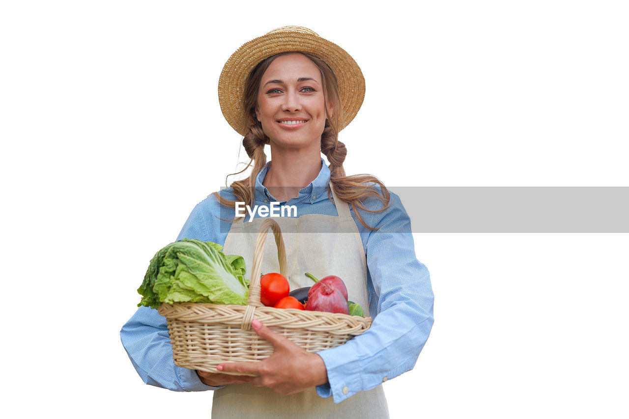 PORTRAIT OF SMILING YOUNG WOMAN HOLDING BASKET OVER WHITE BACKGROUND