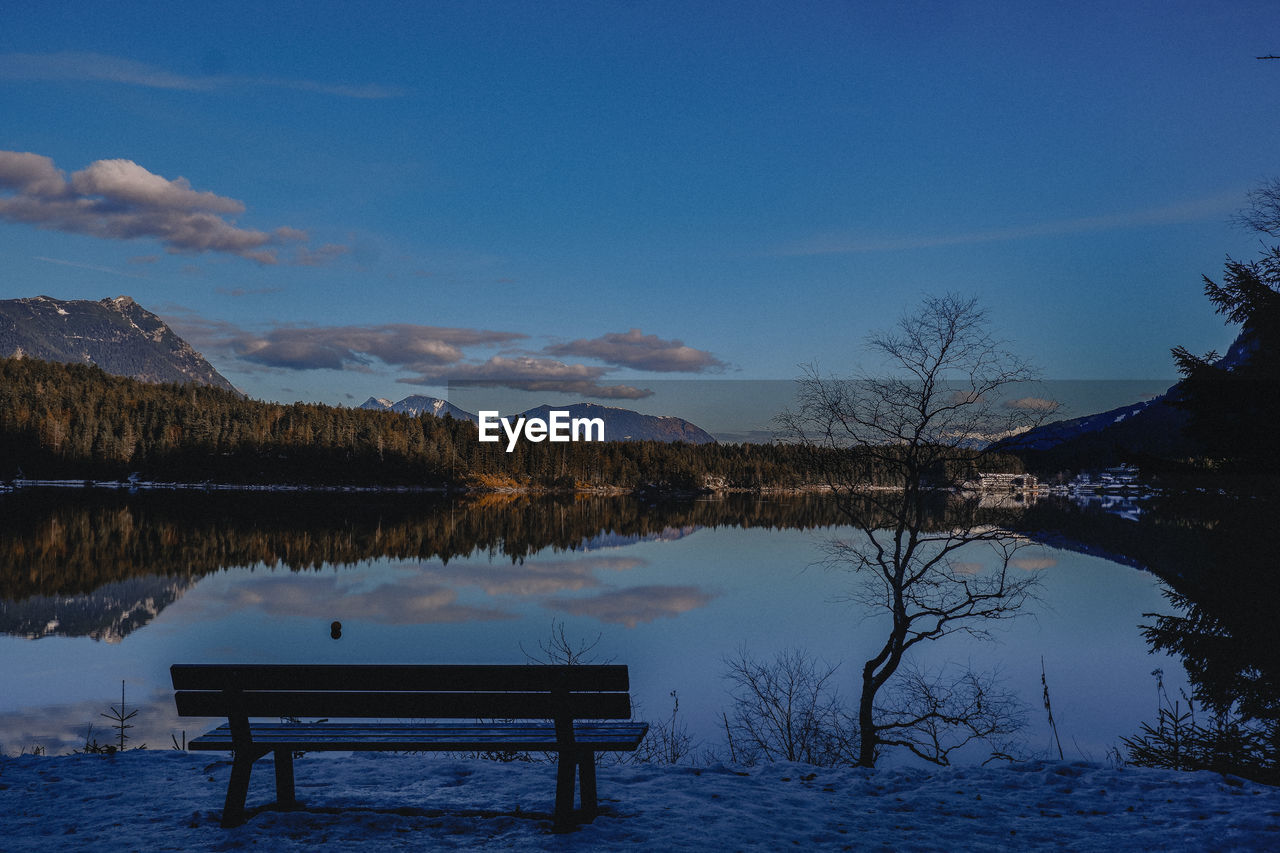 Bench by lake against blue sky during winter