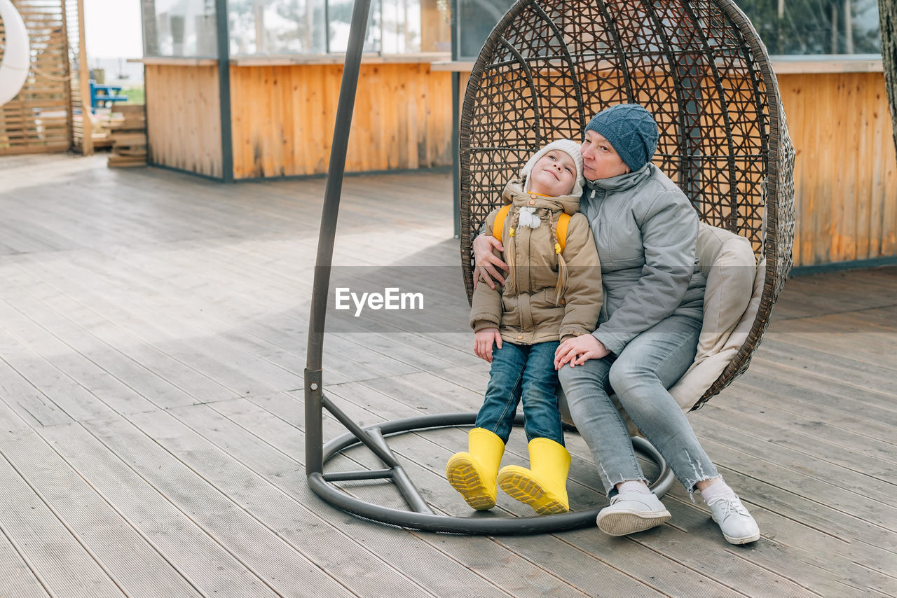 Grandmother and granddaughter sat down to rest after a walk in a hanging rattan chair outdoors