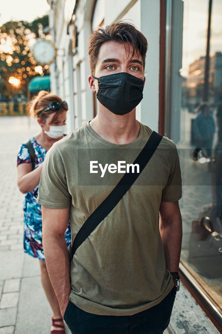 Young man walking along a store front in the city center in the evening wearing the face mask