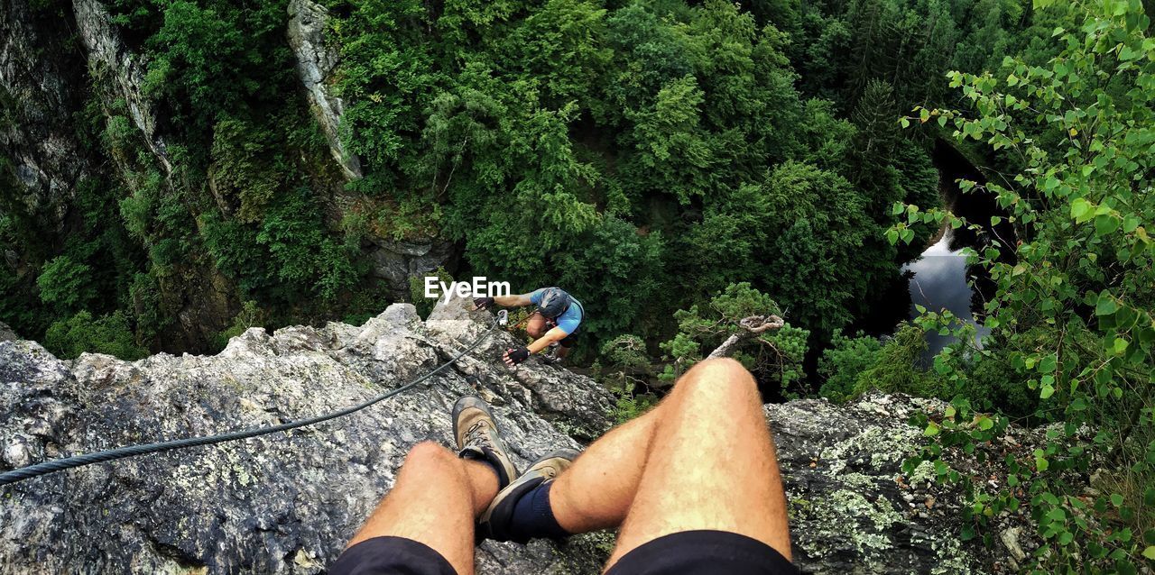 Low section of man sitting on top while person climbing mountain