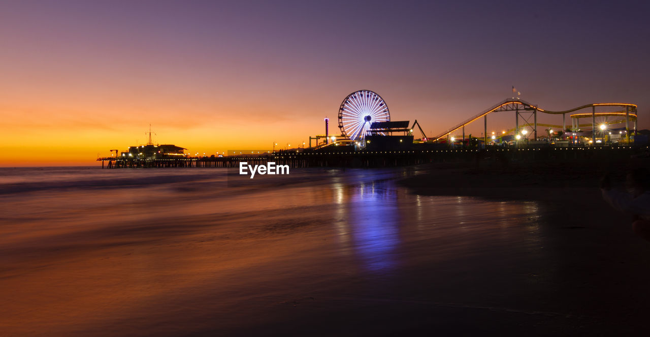 Illuminated amusement park by sea against sky at sunset