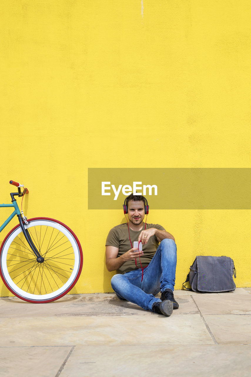 Man listening music while sitting by bicycle on footpath against yellow wall