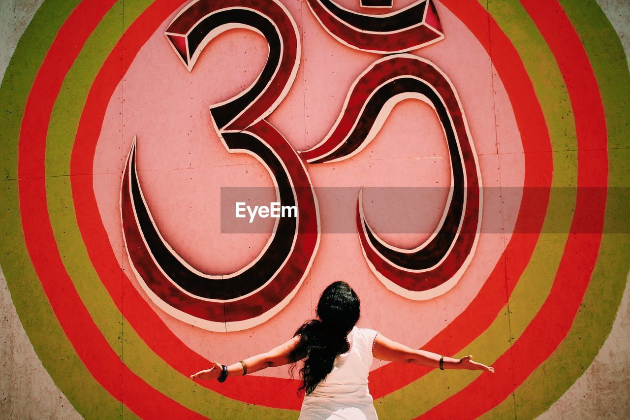 Rear view of woman with arms outstretched standing in front of om symbol on wall