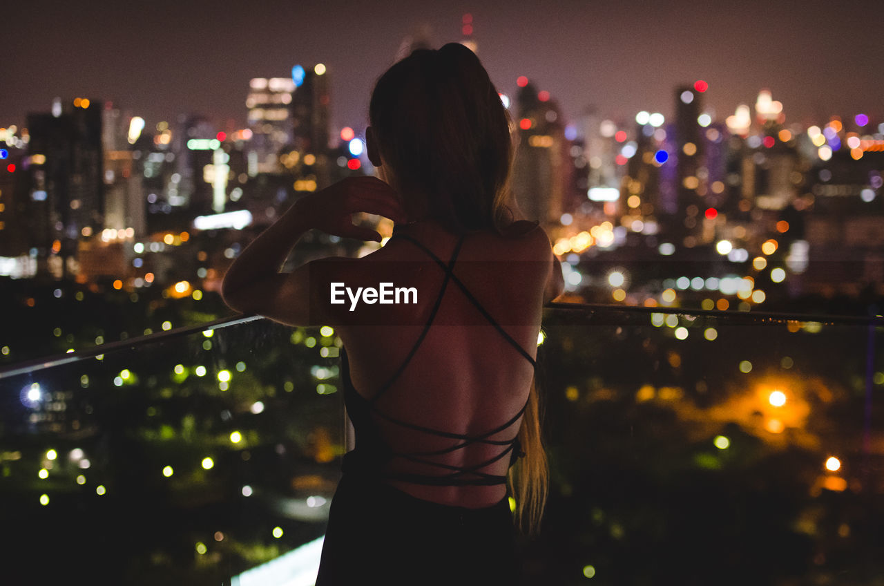 Rear view of woman looking at illuminated cityscape while standing on balcony