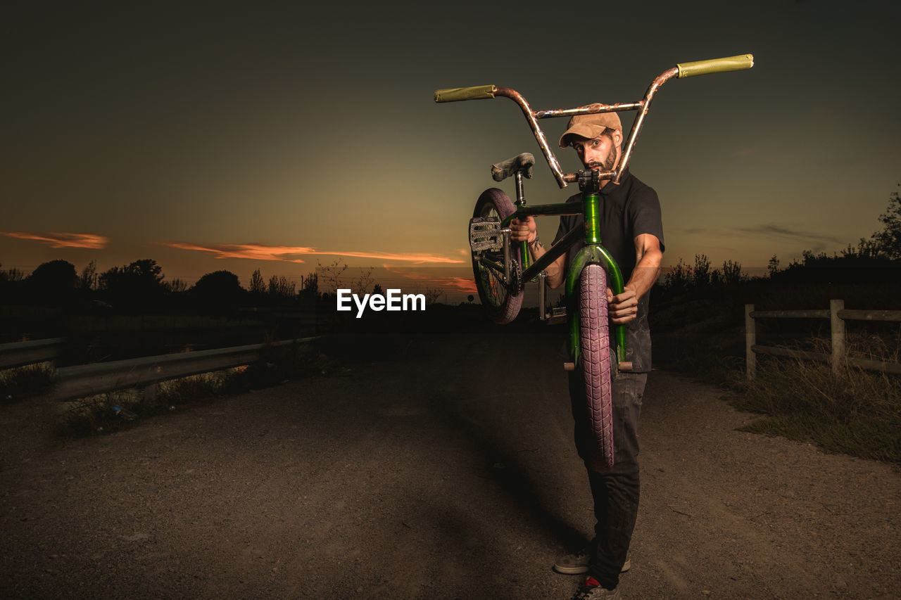 Portrait of young man carrying bicycle while standing on dirt road against sky during sunset