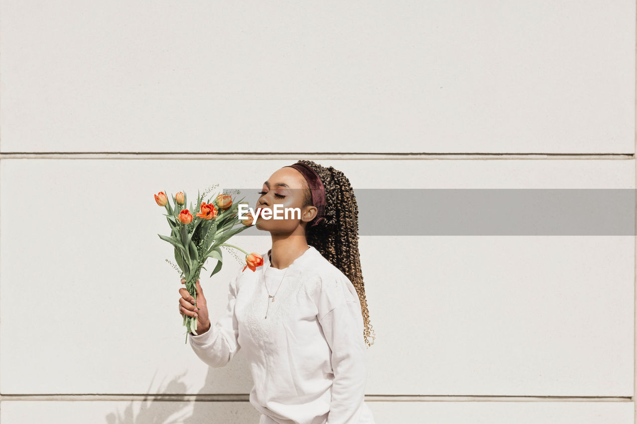 Young woman with eyes closed smelling flowers in front of white wall during sunny day