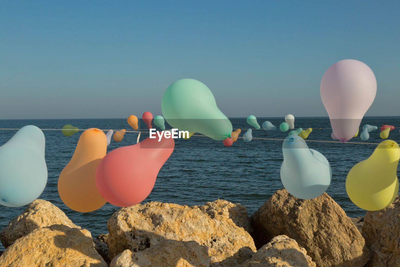 Balloons hanging on rope over rocks against sea and sky during sunny day