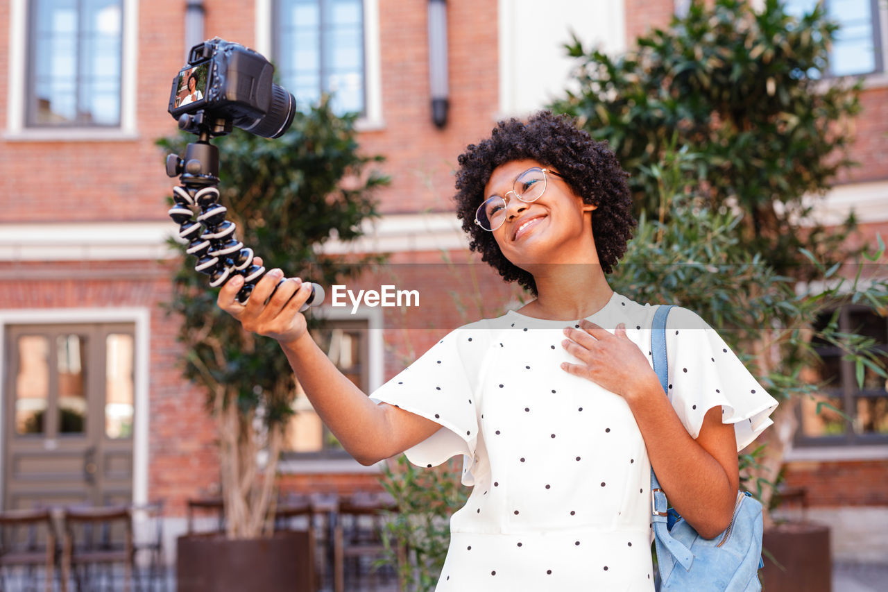 Smiling teenage girl taking selfie with camera while standing against building