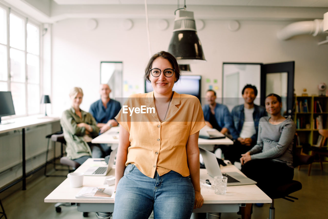 Portrait of smiling female professional leaning on table against executives in modern office