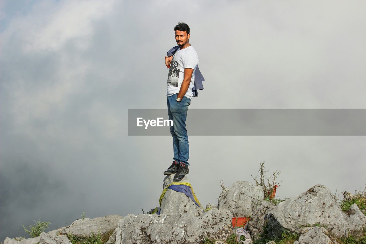 Young man standing on top of rock against cloudy sky