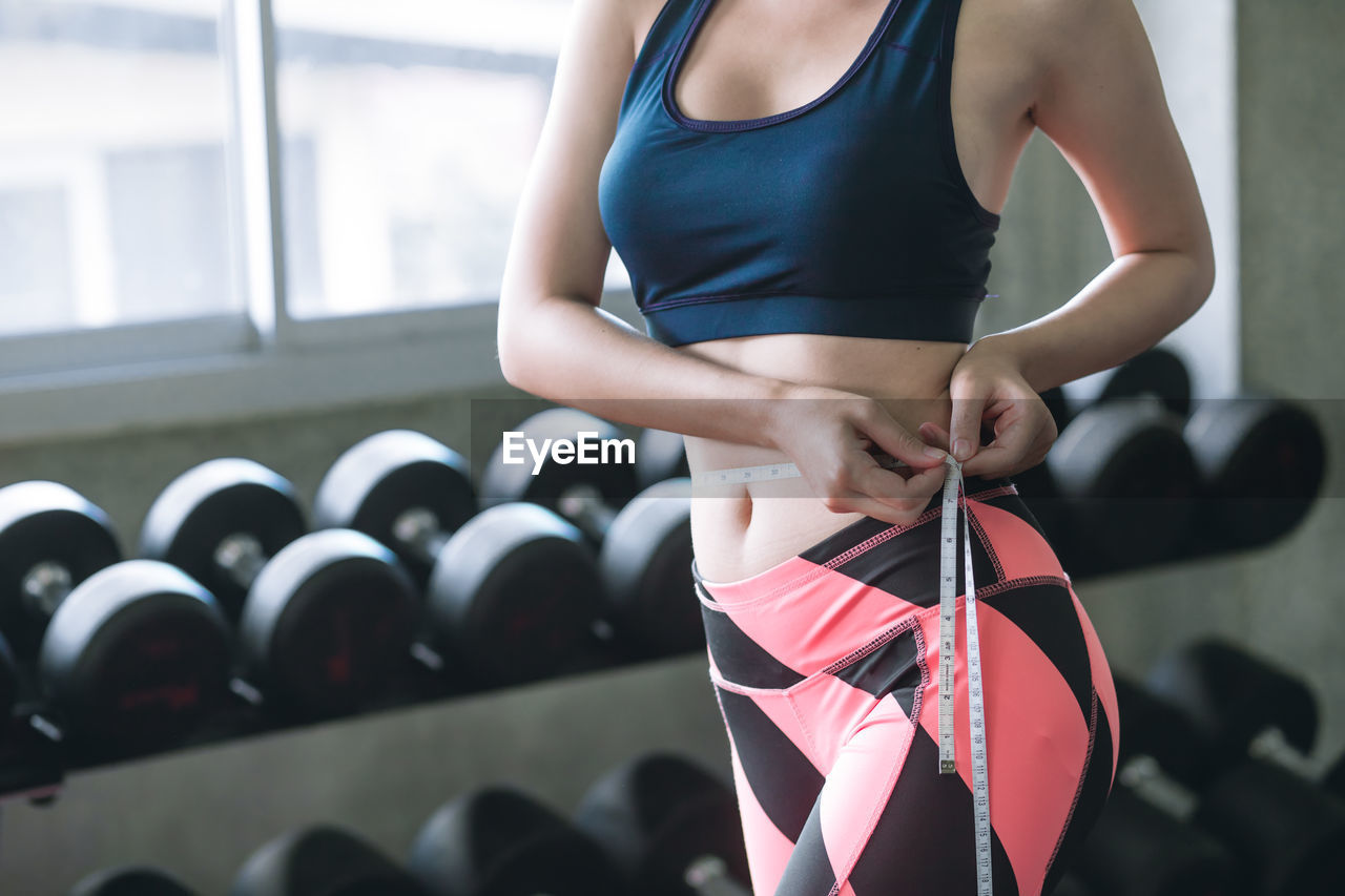 Midsection of woman measuring waist while standing in gym