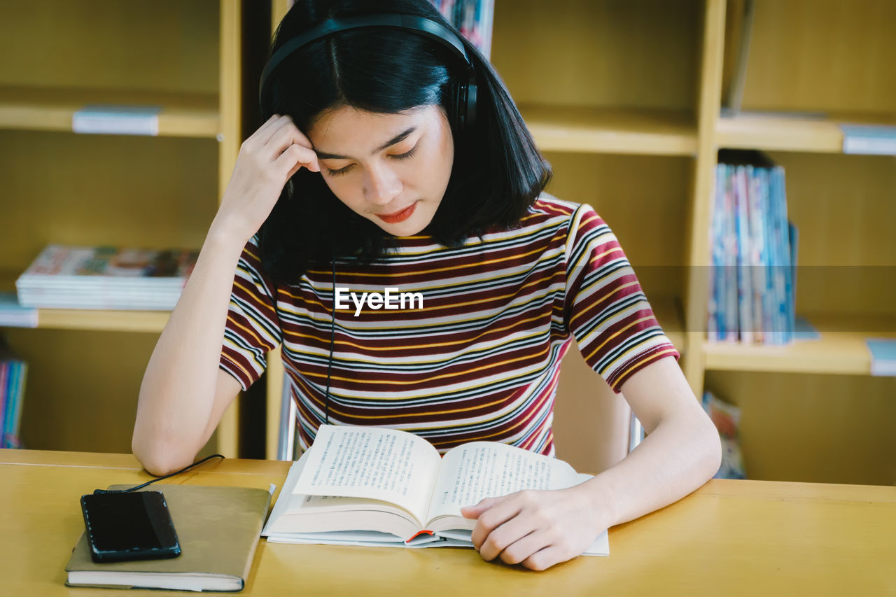 Young woman reading book while listening music on table