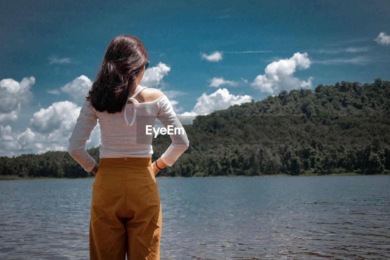 Rear view of woman standing by lake against cloudy sky