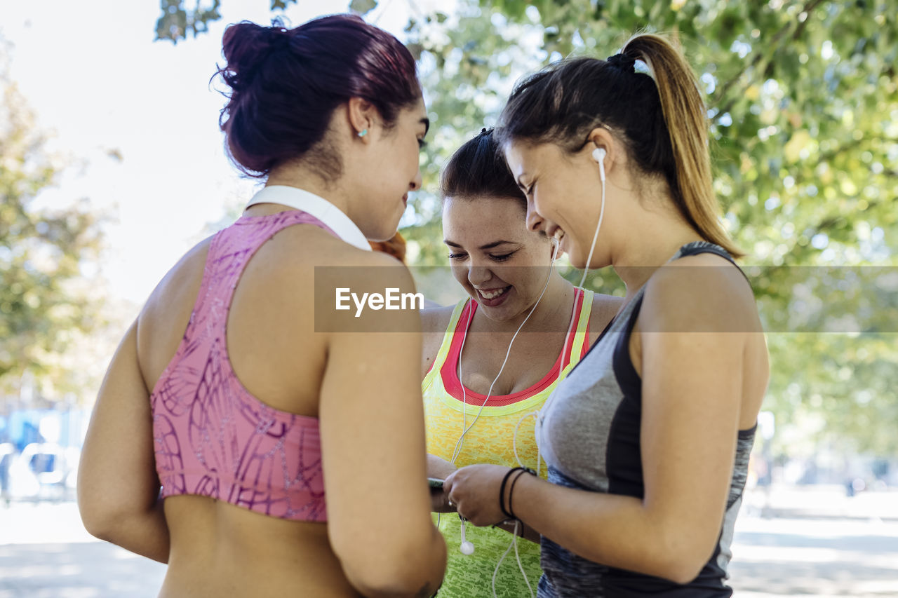 Smiling young woman in sportswear showing mobile phone to female friends at park