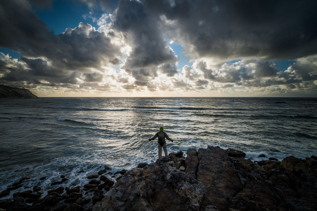 Full length of man standing on rocks at beach against sky during sunset