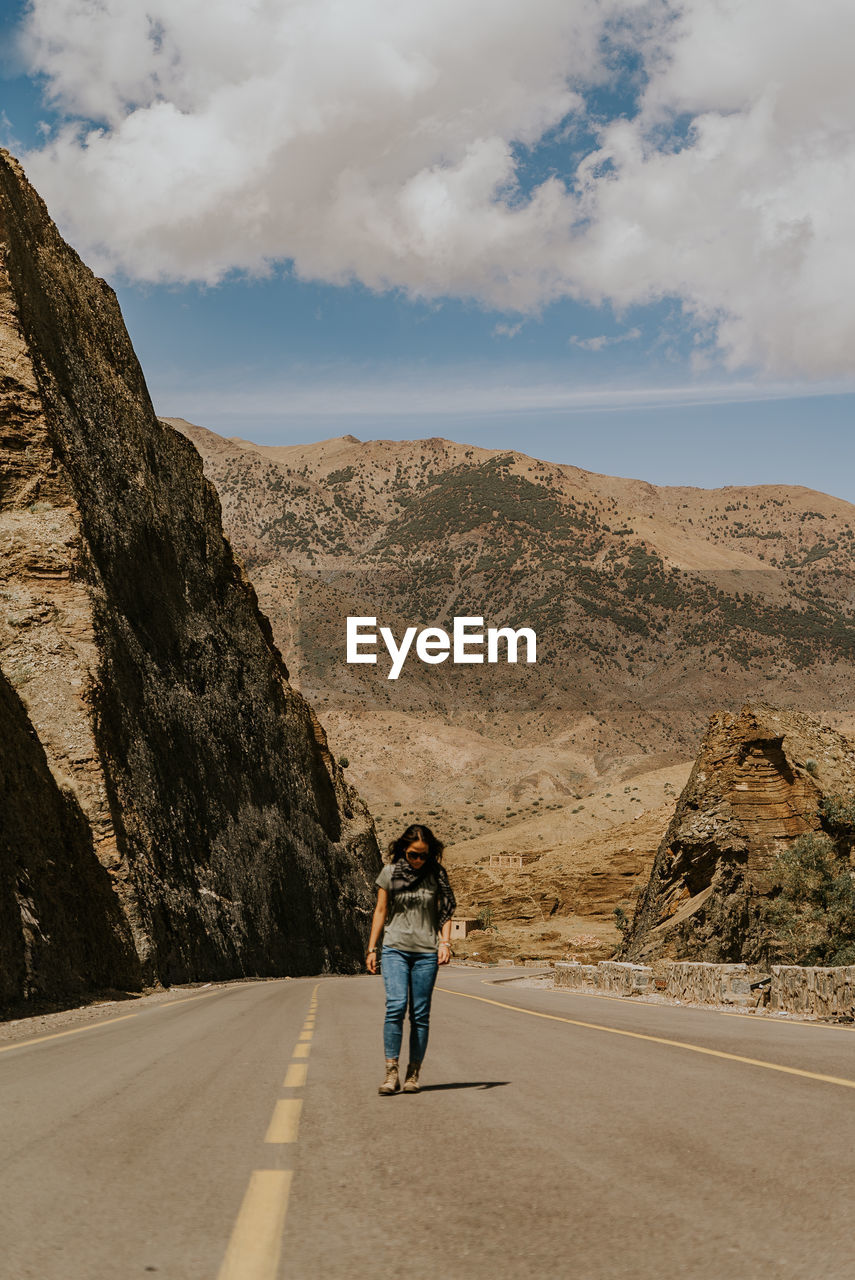 FULL LENGTH REAR VIEW OF WOMAN WALKING ON ROAD AGAINST SKY