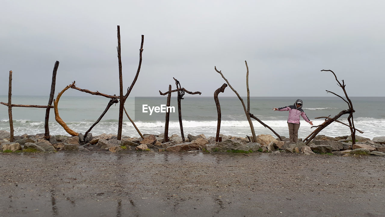 Woman with arms outstretched standing amidst driftwood at beach against sky