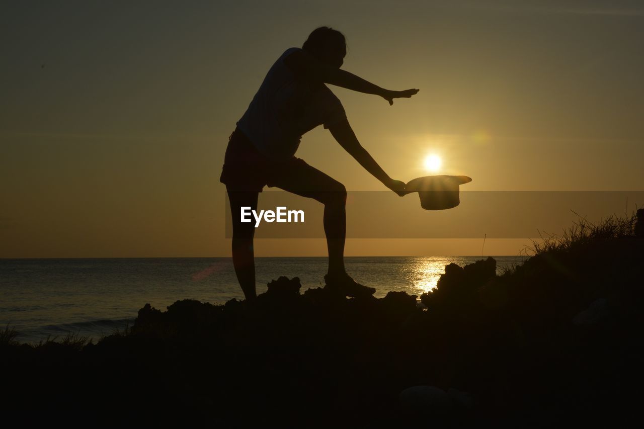 Optical illusion of man putting sun in hat while standing at beach during sunset