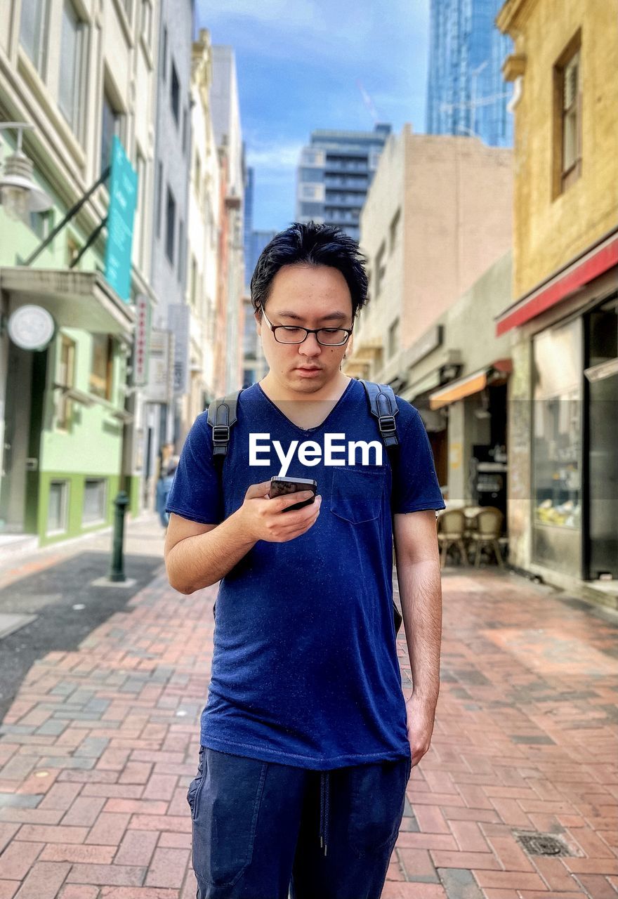 Portrait of young asian man using smartphone while standing in laneway against buildings in city.