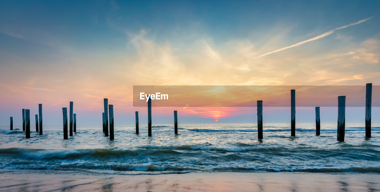 Wooden posts in sea against sky during sunset